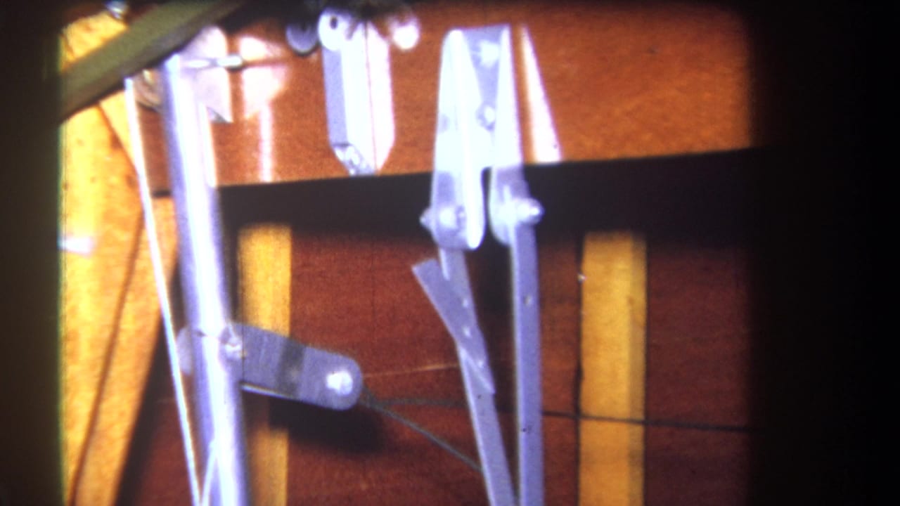 Close 1964 home-movie view of exposed metal linkage, rods, and pivots against a wood-grain cabin or vehicle interior surface.