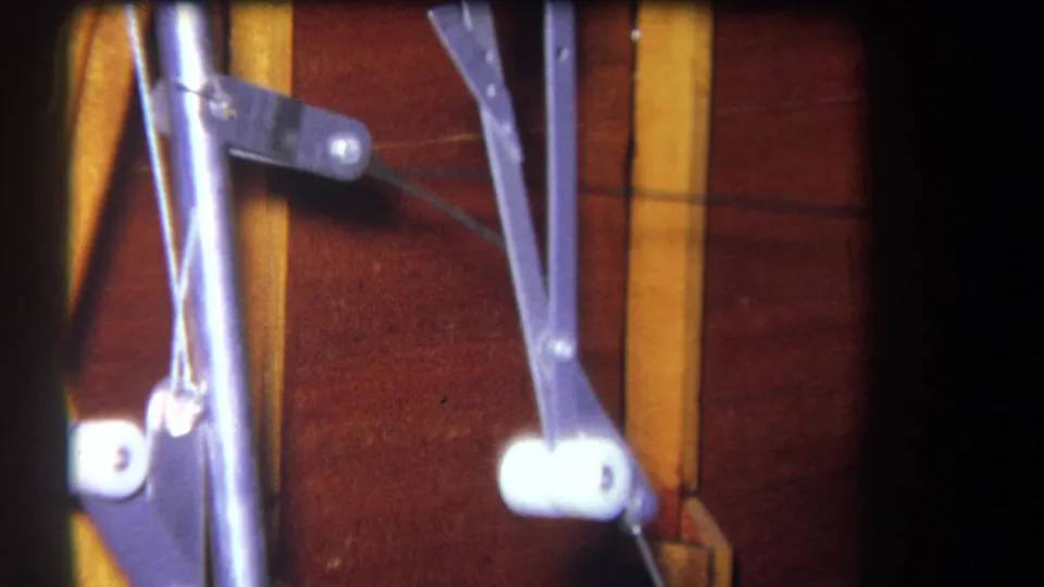 Close 1964 frame of metal control linkage and pivot points beside vertical wood-grain trim, with dark 8mm frame borders visible.