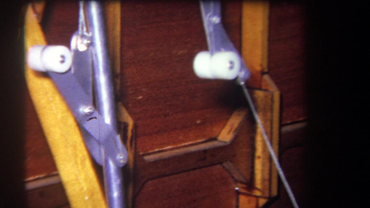 Close 8mm frame of a purple metal mechanism with white rollers and a cable running beside a wooden support.