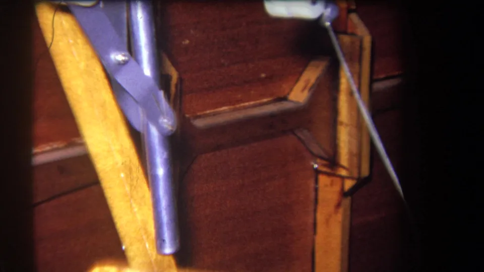 Final close 8mm frame of the mechanical pulley area, with a white roller, cable, purple metal arm, and wood panels.