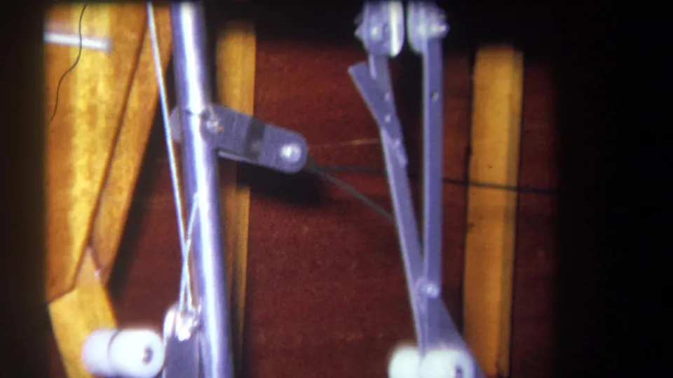 1964 home-movie frame showing a closer view of metal arms, rods, a white roller, and yellow wooden supports.