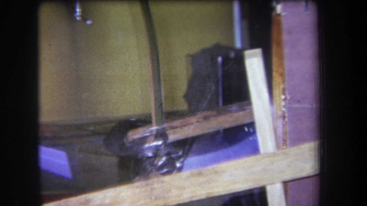 1964 Yucaipa-area home-movie frame showing a curved wood support, purple work surface, black cable, clamp hardware, and upright boards in a workshop setting.