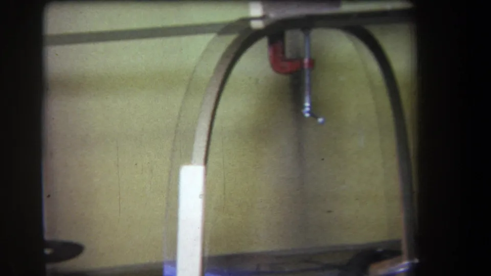 1964 Yucaipa-area home-movie frame showing a simple curved wooden frame with a red clamp attached, cables below, and yellow wall panels behind it.