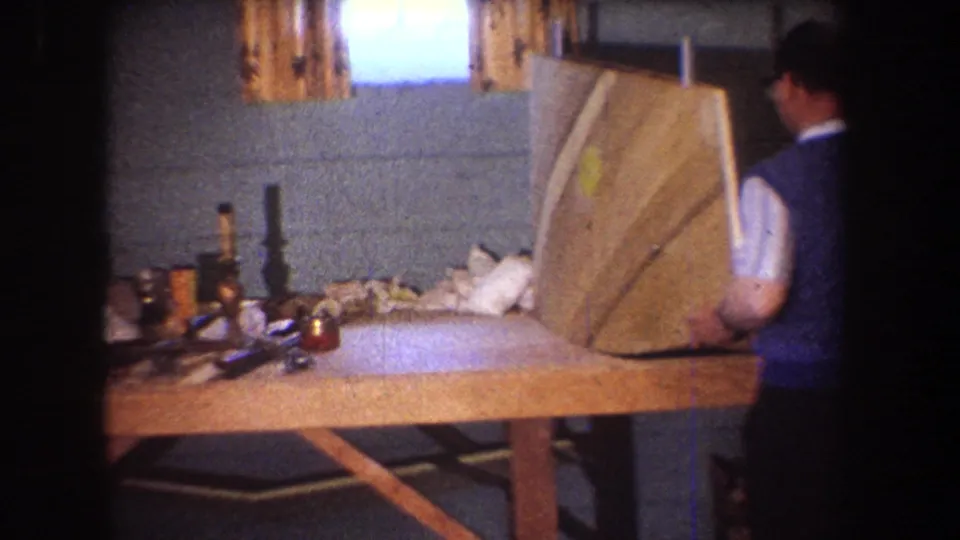 A man in a blue vest works with a large wooden panel at a cluttered workbench in a private 1965 home-movie interior.