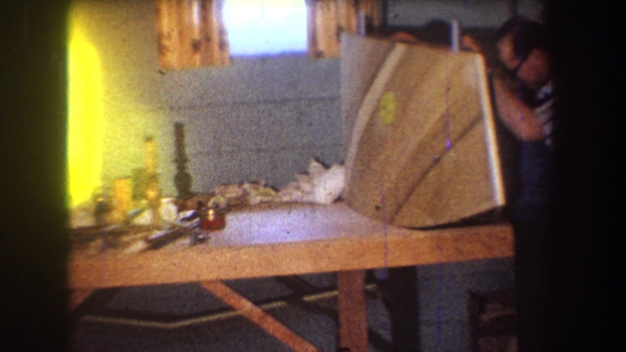 Man beside a workbench lifts or positions a curved wooden panel in a small workshop.