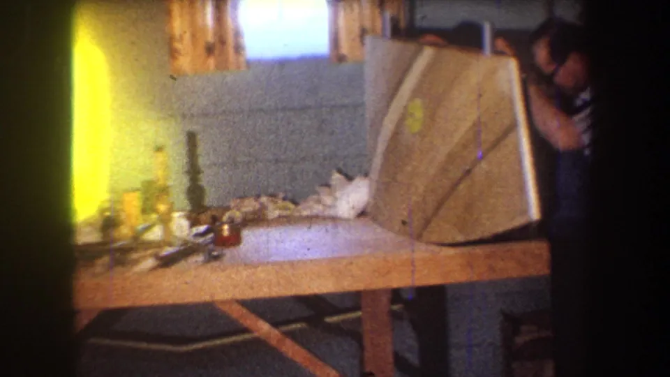 1965 Yucaipa home-movie frame showing a man in a workshop handling a large curved wooden panel on a cluttered bench.