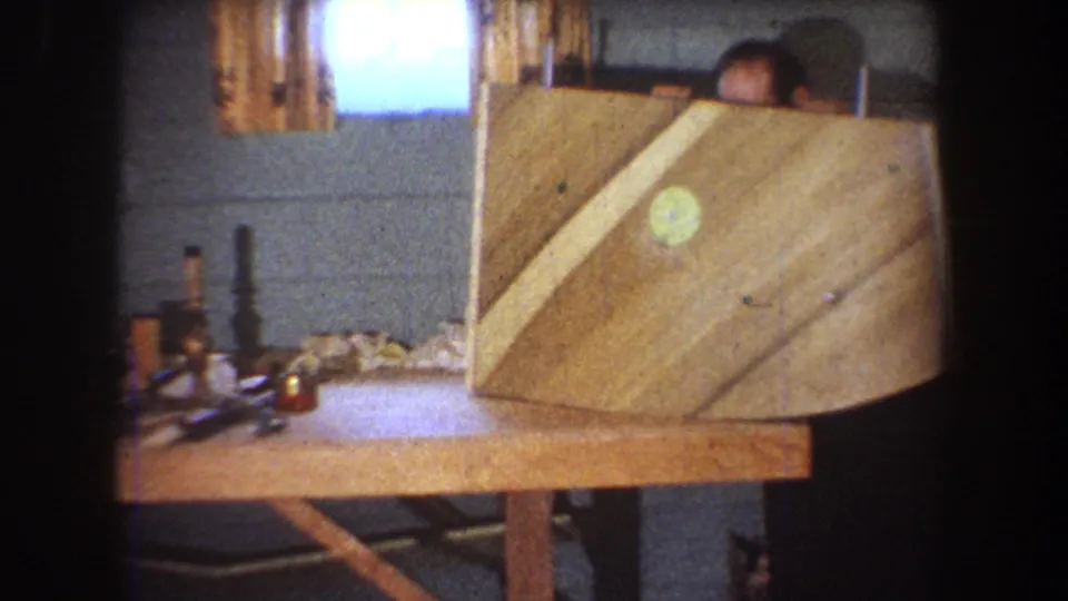 Home-movie frame from 1965 Yucaipa showing a large curved wooden panel dominating a workbench as a man appears behind it.