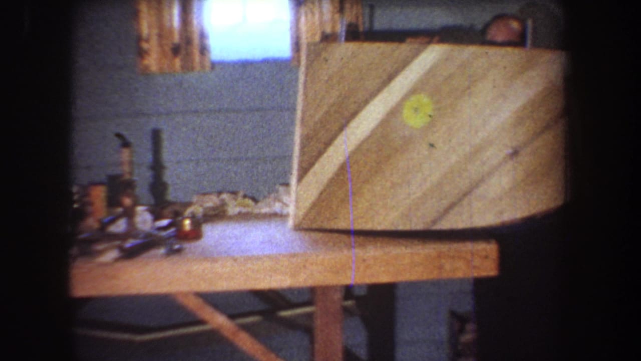 In a 1965 Yucaipa home workshop, a wooden boat hull sits upright on a bench near tools and a curtained window.