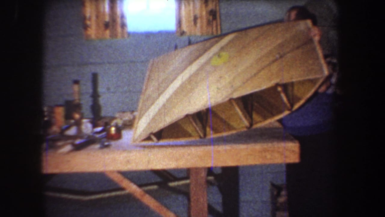 A man begins lifting a curved wooden boat hull from the workbench.