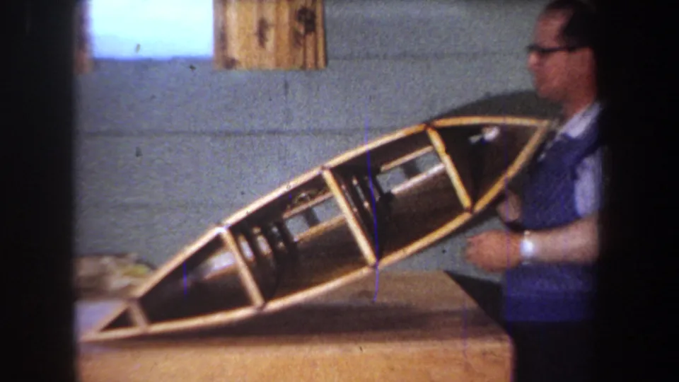 A man in glasses stands beside the angled handmade hull, with its internal framing visible on the workbench.