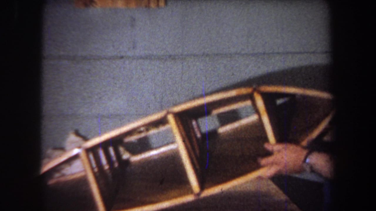 1965 home-movie frame from Yucaipa showing hands holding a wooden model boat in a close handheld view.