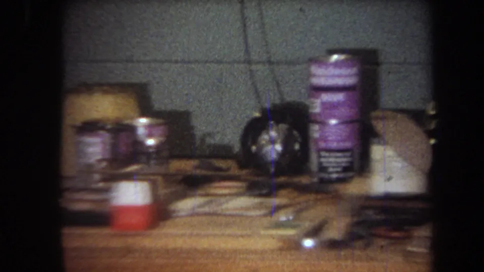 1965 Yucaipa-area workshop table showing cans, a dark object, cords, tools, and small craft materials in soft 8mm focus.