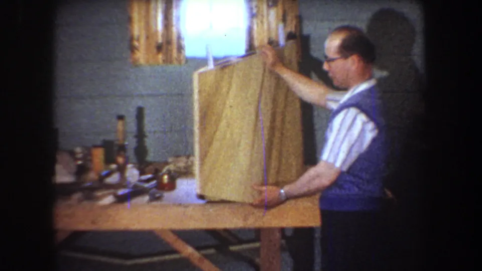In the 1965 Yucaipa home-movie frame, a man in glasses and a blue vest positions a large board on a basement workbench.