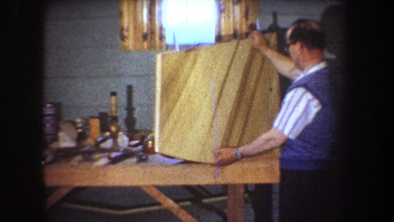 Man in a blue vest holds a large wooden panel on a basement workbench.