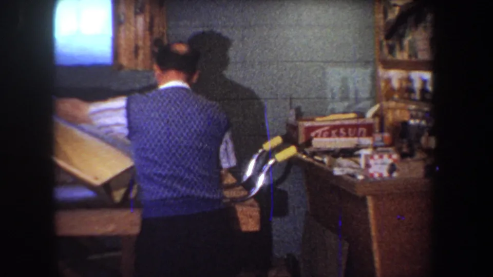 1965 Yucaipa home workshop frame showing the man moving a large wooden panel with shelves and supplies nearby.