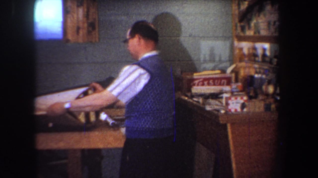 In 1965 home-movie footage from Yucaipa, California, a man in glasses and a blue sweater vest handles a frame or fixture at a wooden worktable.