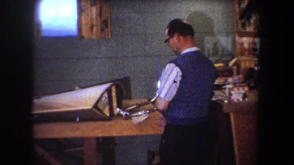 Seen from behind, the man works at the table with a triangular frame or fixture and nearby tools in a private basement workshop.