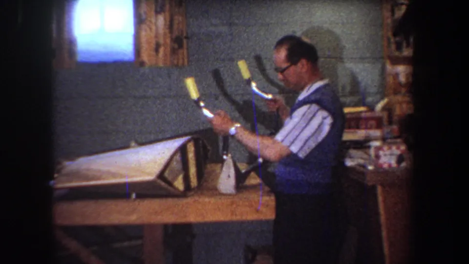 The man holds up a metal fork-shaped bicycle part or tool while standing beside the worktable in the 1965 basement workshop scene.
