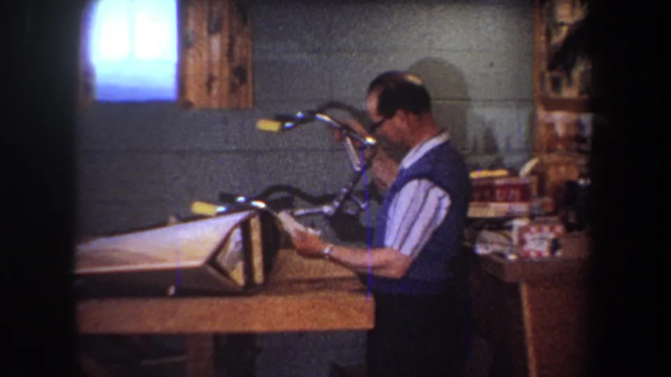 The man continues examining the bicycle handlebars in a private workshop, with yellow grips and a small window visible.