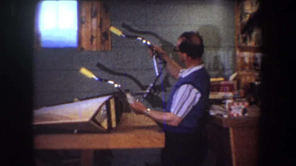 A man in a blue vest lifts or steadies the bicycle handlebars for inspection in the 1965 Yucaipa workshop scene.