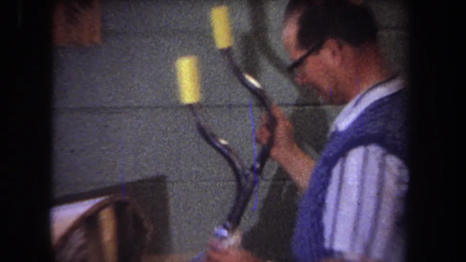 A closer 8mm frame shows the man in glasses focused on curved bicycle handlebars with yellow grips inside the workshop.