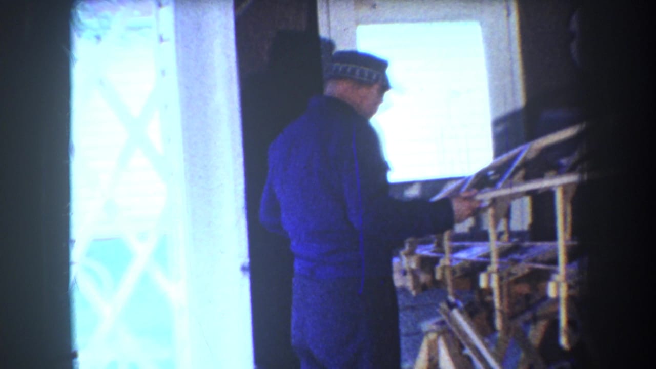 Man in blue work clothes stands beside a wooden aircraft frame near a bright window.