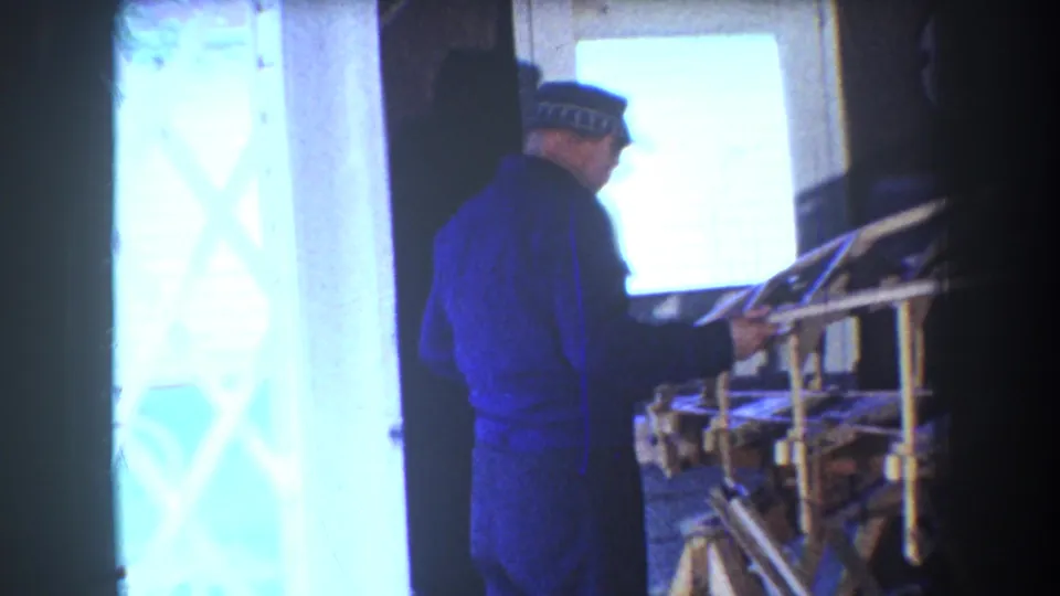 1965 Yucaipa home-movie frame showing an adult man in blue work clothes inspecting a wooden airplane frame inside a workshop.