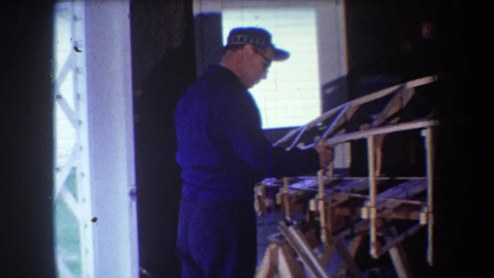 1965 Yucaipa 8mm frame showing an adult man in a cap handling part of a wooden airplane frame in a dim workshop.