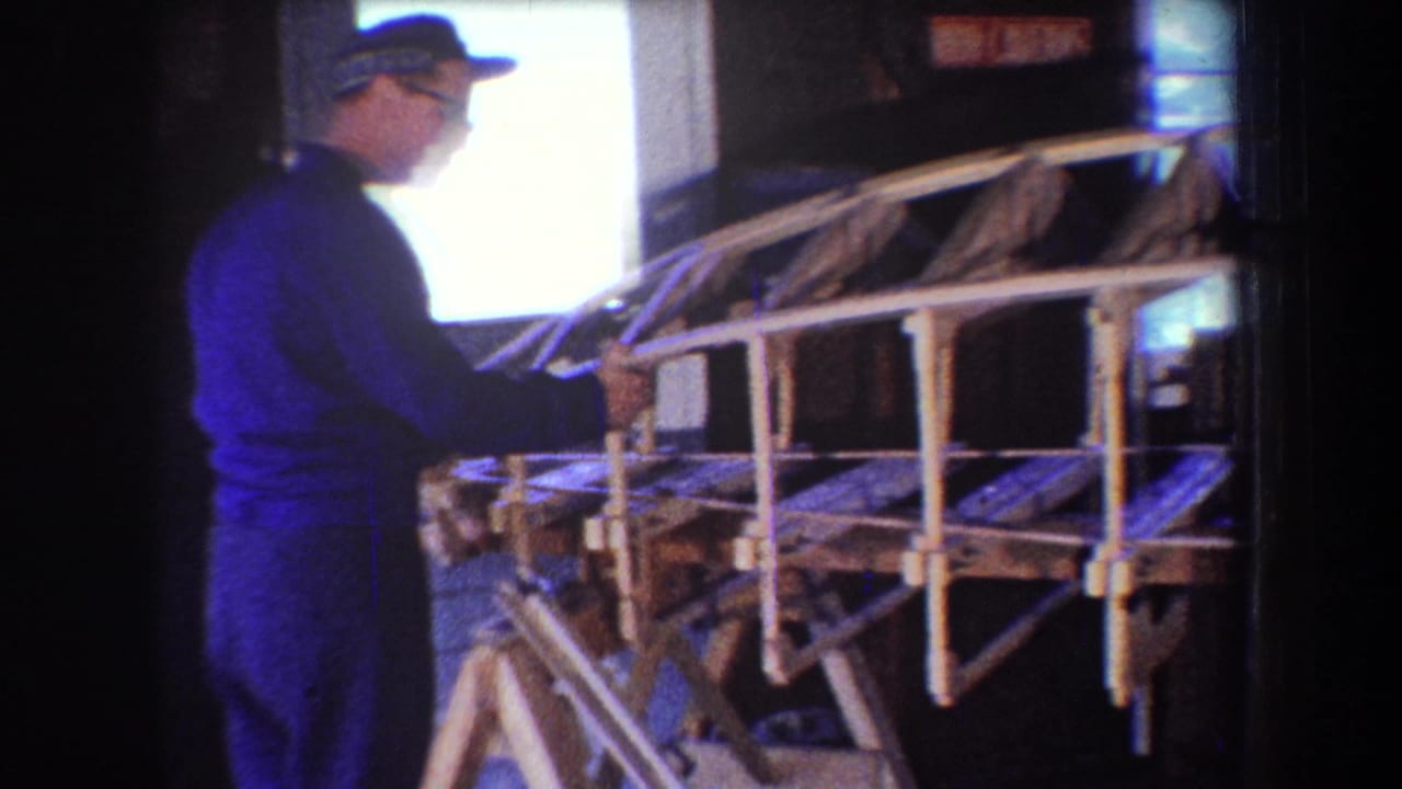 Man in a cap stands beside a large wooden frame inside a dim workshop.