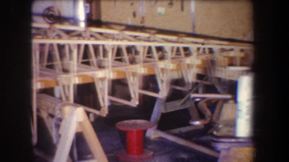 A wooden aircraft frame extends across a 1965 private workshop view, with a red spool and equipment on the floor nearby.