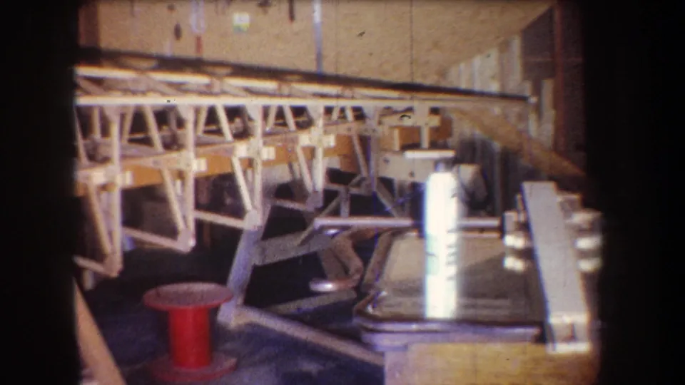 The camera looks along the unfinished wooden aircraft frame, showing supports, a red spool, and workshop items in the foreground.