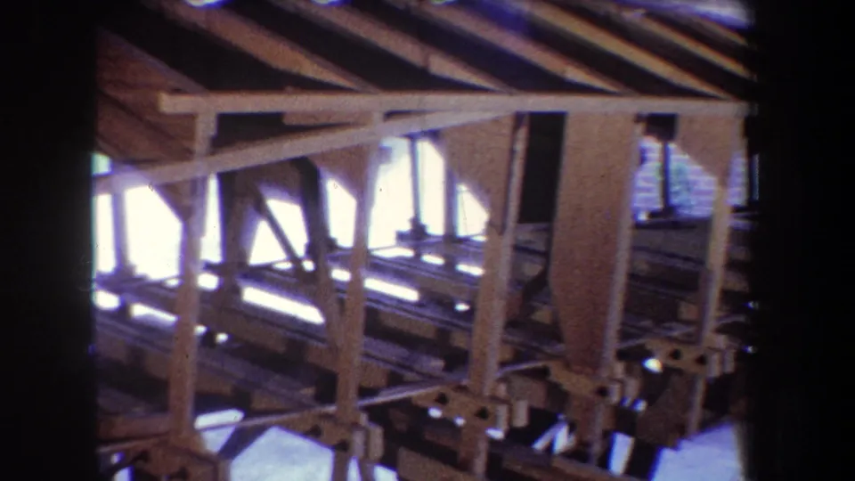 1965 8mm home-movie frame from Yucaipa, California showing exposed beams, vertical supports, rafters, and bright exterior light through an unfinished wooden structure.