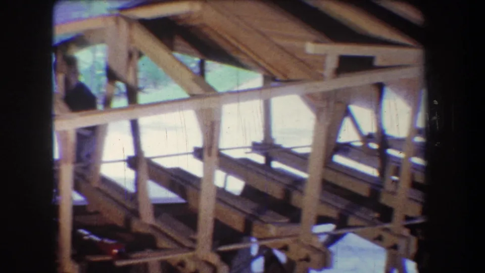 1965 Yucaipa construction footage showing a partially framed wooden interior with rafters, diagonal braces, wall openings, and a worker briefly visible in dark clothing.