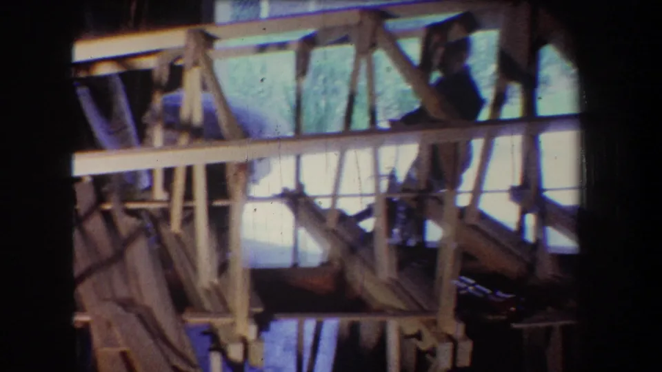 8mm frame from the 1965 Yucaipa clip showing dense wooden framing, diagonal braces, open wall sections, and a partially obscured worker within the construction area.