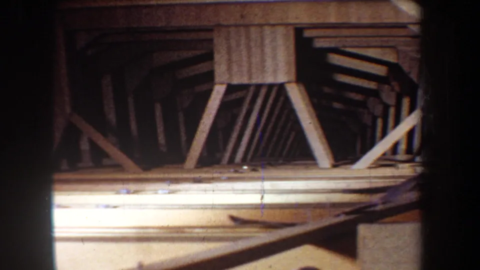 1965 Yucaipa 8mm frame showing repeated timber framing and diagonal supports inside a shadowed construction area.