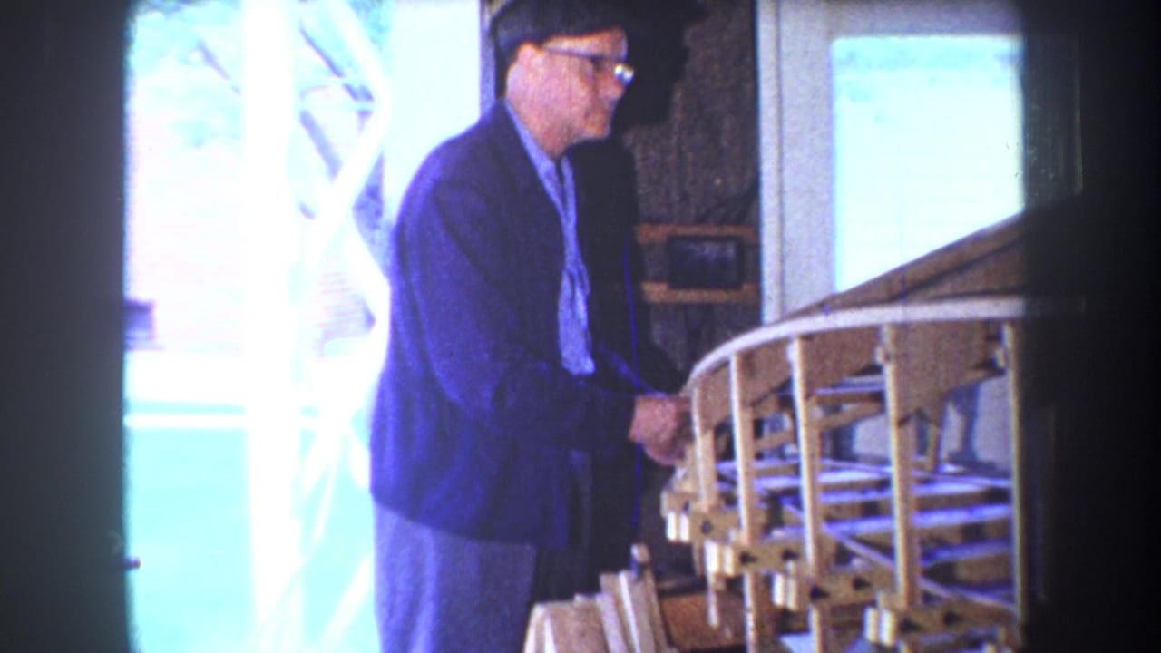 In 1965 Yucaipa home-movie footage, a man in glasses stands near a wooden model airplane frame by a porch railing.