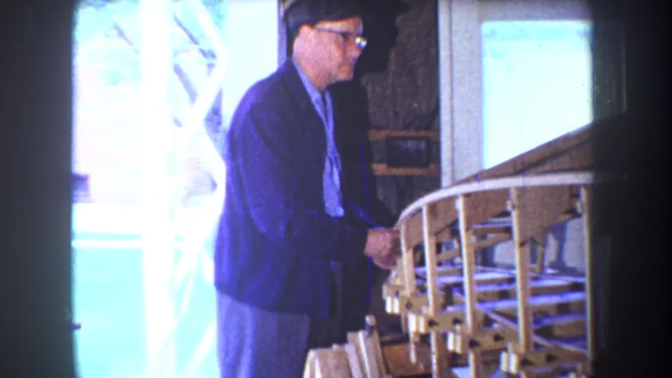 In 1965 Yucaipa home-movie footage, a man in glasses stands near a wooden model airplane frame by a porch railing.