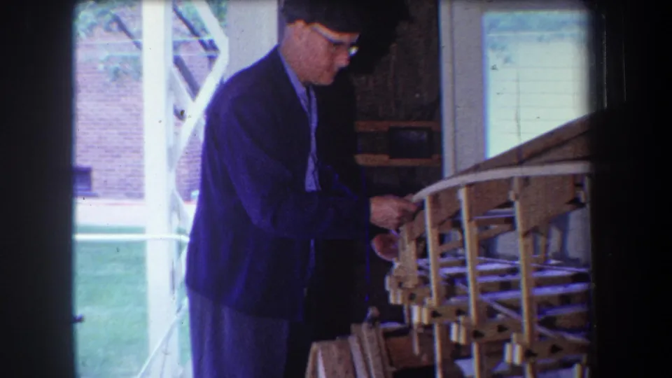 A man in glasses adjusts a wooden model airplane frame on a porch in supported 1965 Yucaipa home-movie footage.
