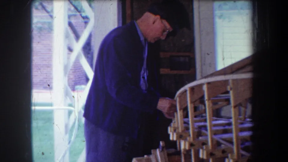 The 1965 8mm frame shows the man leaning over the wooden airplane structure with a bright porch opening behind him.