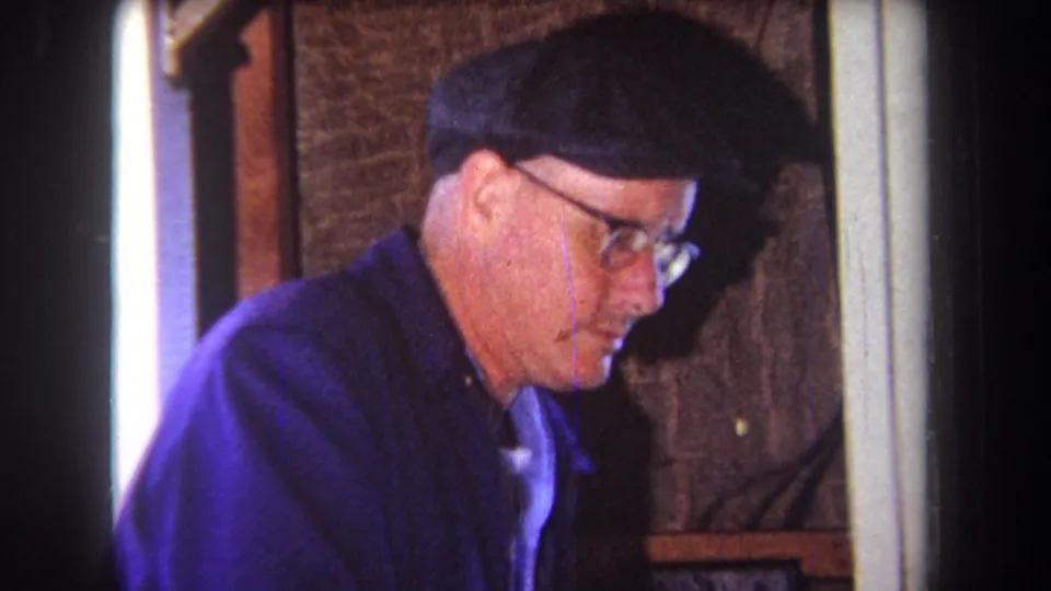 A close 8mm frame shows the man's face, cap, glasses, and blue work jacket as he focuses on the woodworking area.