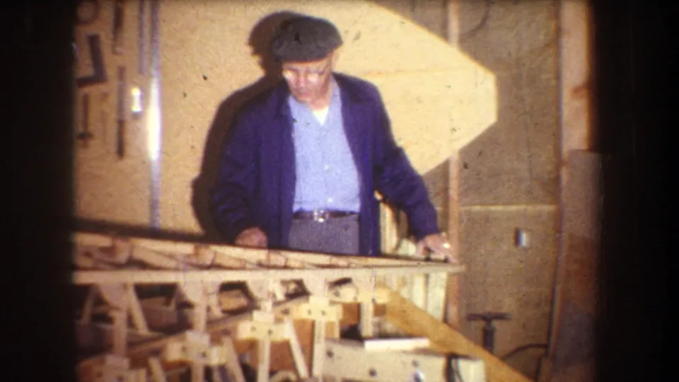 The man stands behind a partly built wooden structure, with tools and workshop surfaces visible in the private interior.