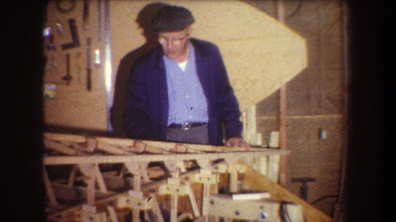 Older man in a flat cap stands behind a wooden frame on a workbench.
