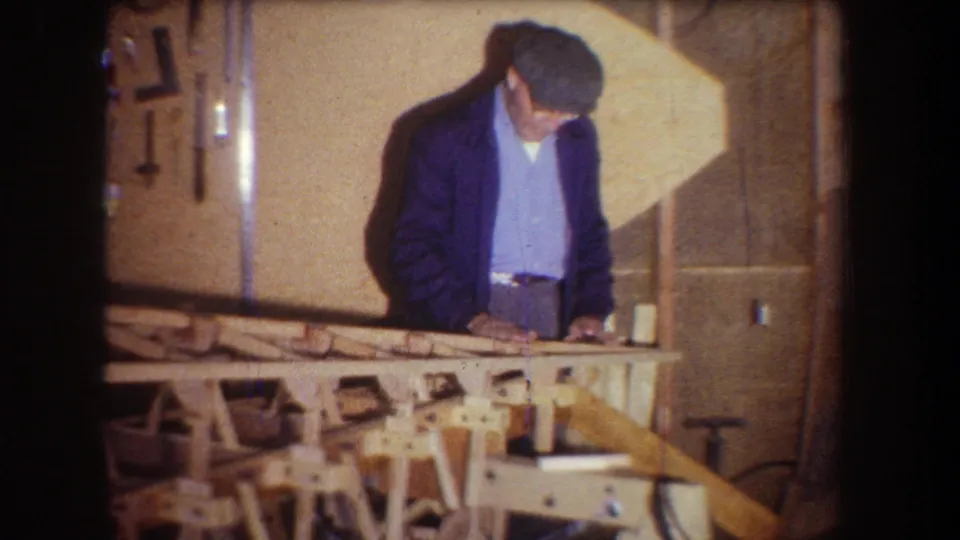 In a 1965 workshop scene, the man leans over the wooden frame as tools hang on the wall behind him.