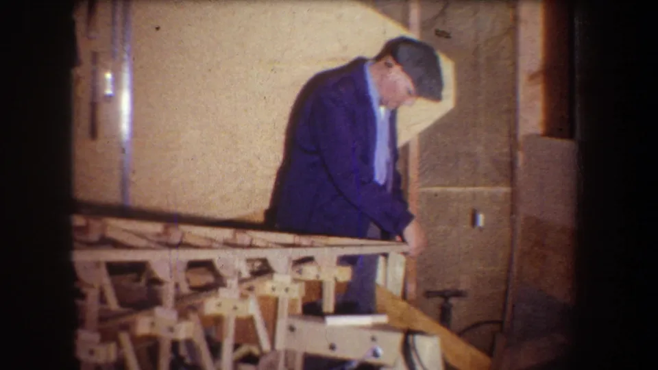 The older man works near the end of a wooden frame inside a private workshop with warm 8mm film color.