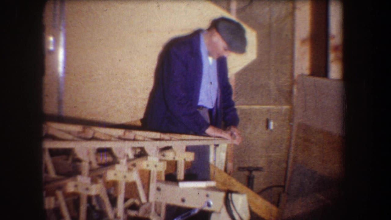 Man in a flat cap leans over a wooden model on a workbench indoors.
