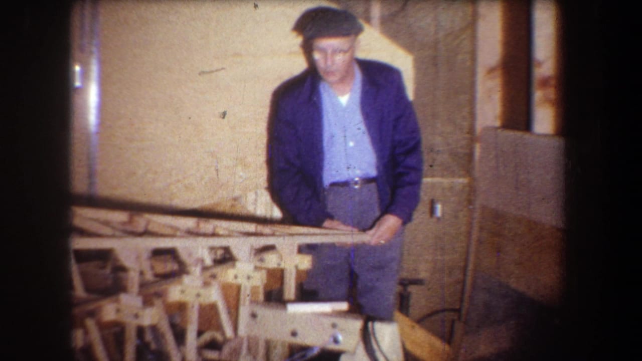 1965 Volmer Yucaipa frame of a man in a cap, glasses, and blue jacket standing at a workshop bench with wood pieces.