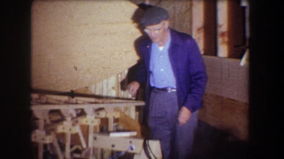 1965 Volmer Yucaipa home-movie frame showing the man stepping near a wooden model and workbench in a private interior.
