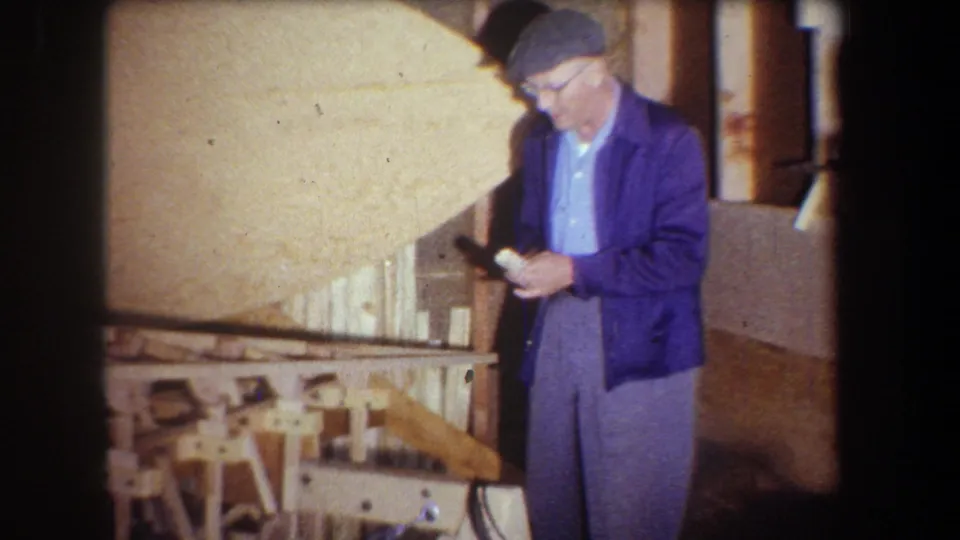 1965 Volmer Yucaipa frame of a man with glasses examining a small item beside a wooden model in a workshop.