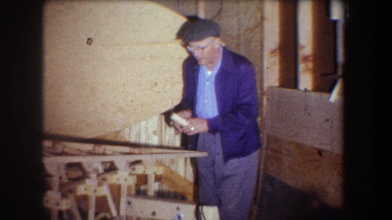 1965 Yucaipa home-movie frame showing a man in a blue jacket and flat cap handling a tool near an unfinished wooden structure.