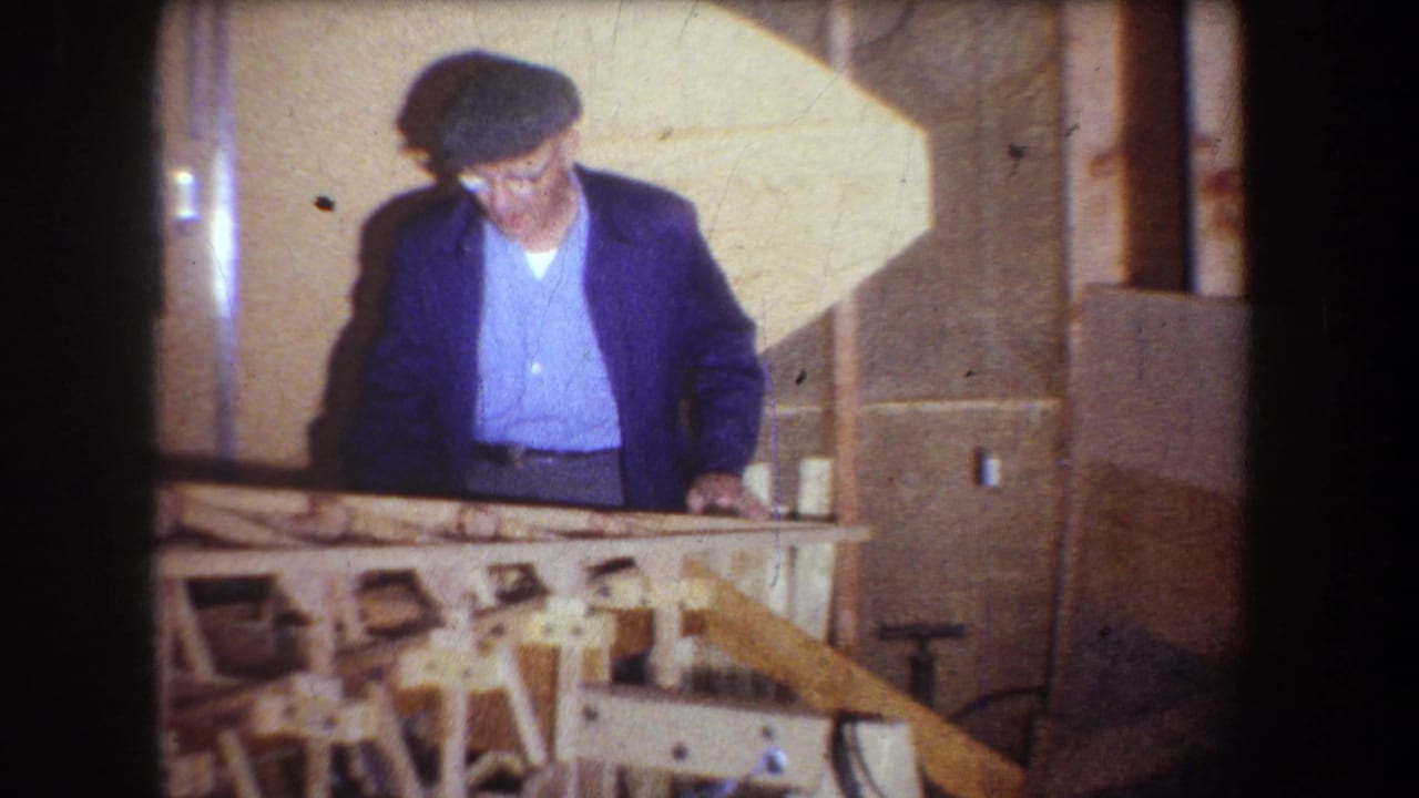 1965 Yucaipa frame with a man in a cap and blue jacket standing over a partly assembled wooden project in an indoor workspace.